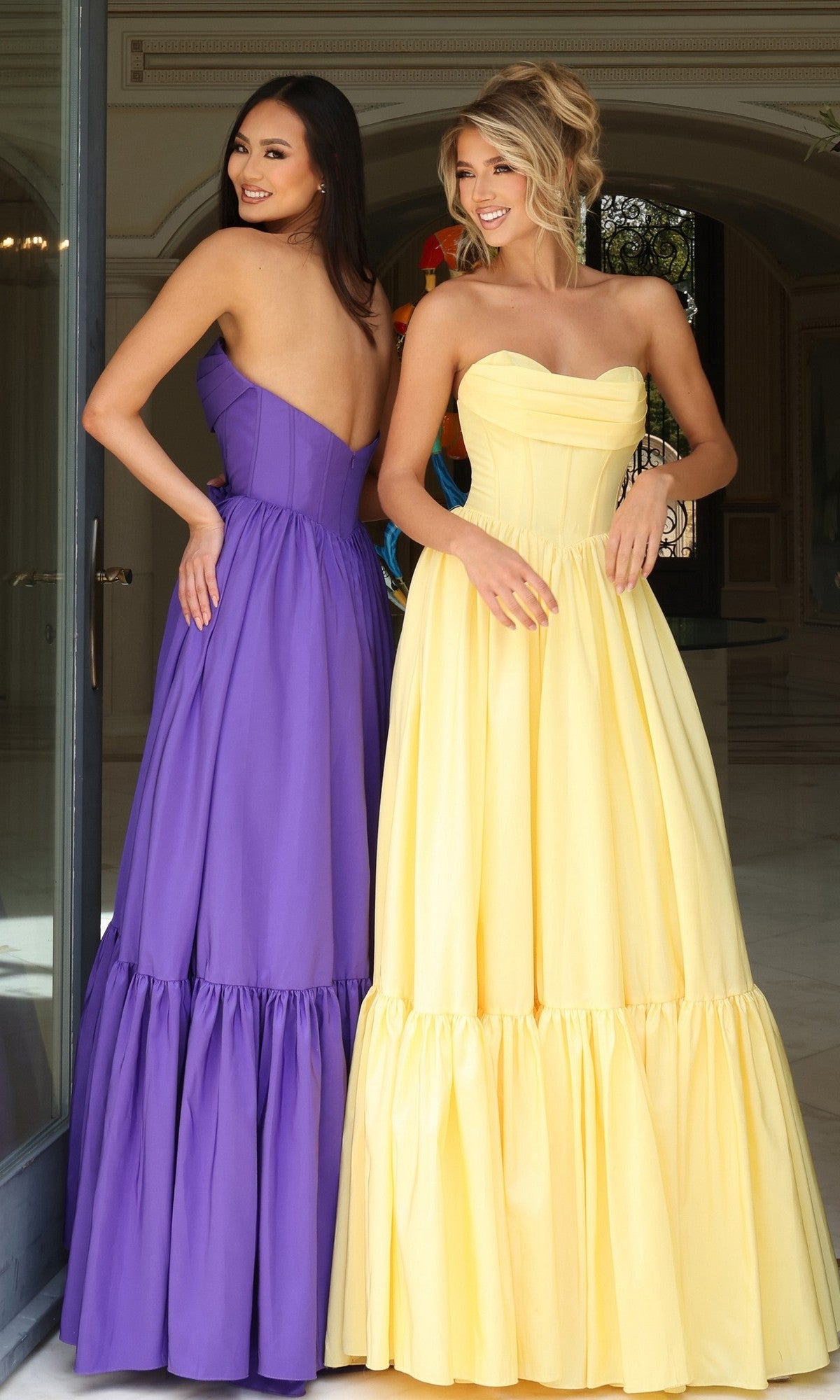 Two women stand indoors, smiling and looking over their shoulders. One wears the Jovani 47883 Strapless Long Ruffled Prom Dress with a corset-style bodice, while the other is in a yellow prom dress.